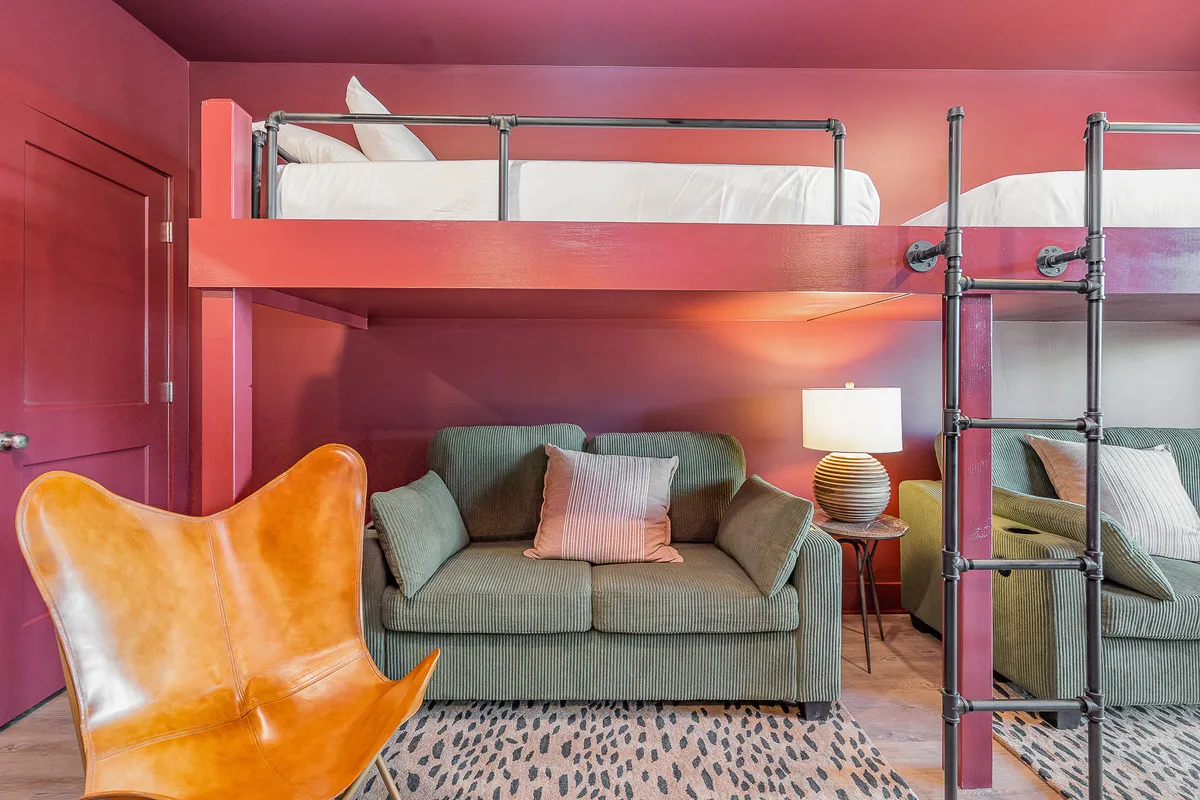 A modern, inviting loft space with a lofted bed, butterfly chair, green sofas, and a table lamp. The bright, open setting offers a mix of seating and sleeping options, with a casual, social feel ideal for group gatherings or quiet downtime.