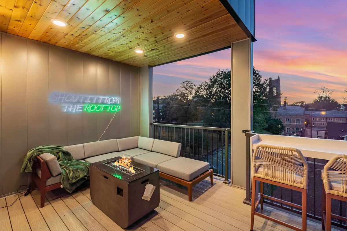 A stylish rooftop outdoor lounge featuring a cozy seating area encircling a fire pit, with a neon sign adorning the space. The seating is oriented to capture a vibrant view of the sunset, creating a welcoming atmosphere for socializing and relaxation.