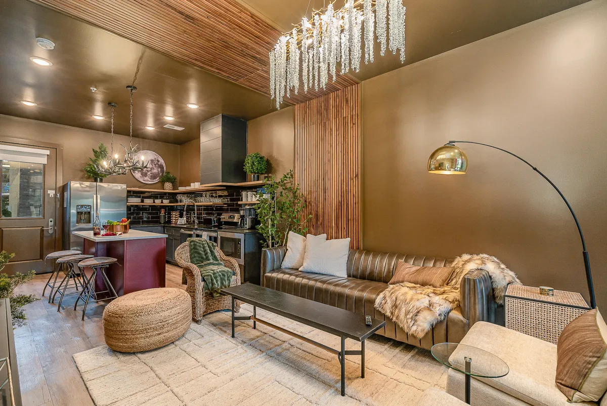A tastefully arranged living area featuring a grand chandelier, a plush sofa, a spacious island bar, and lush indoor plants. The layout suggests a space designed for both social interaction and personal relaxation in a sophisticated setting.