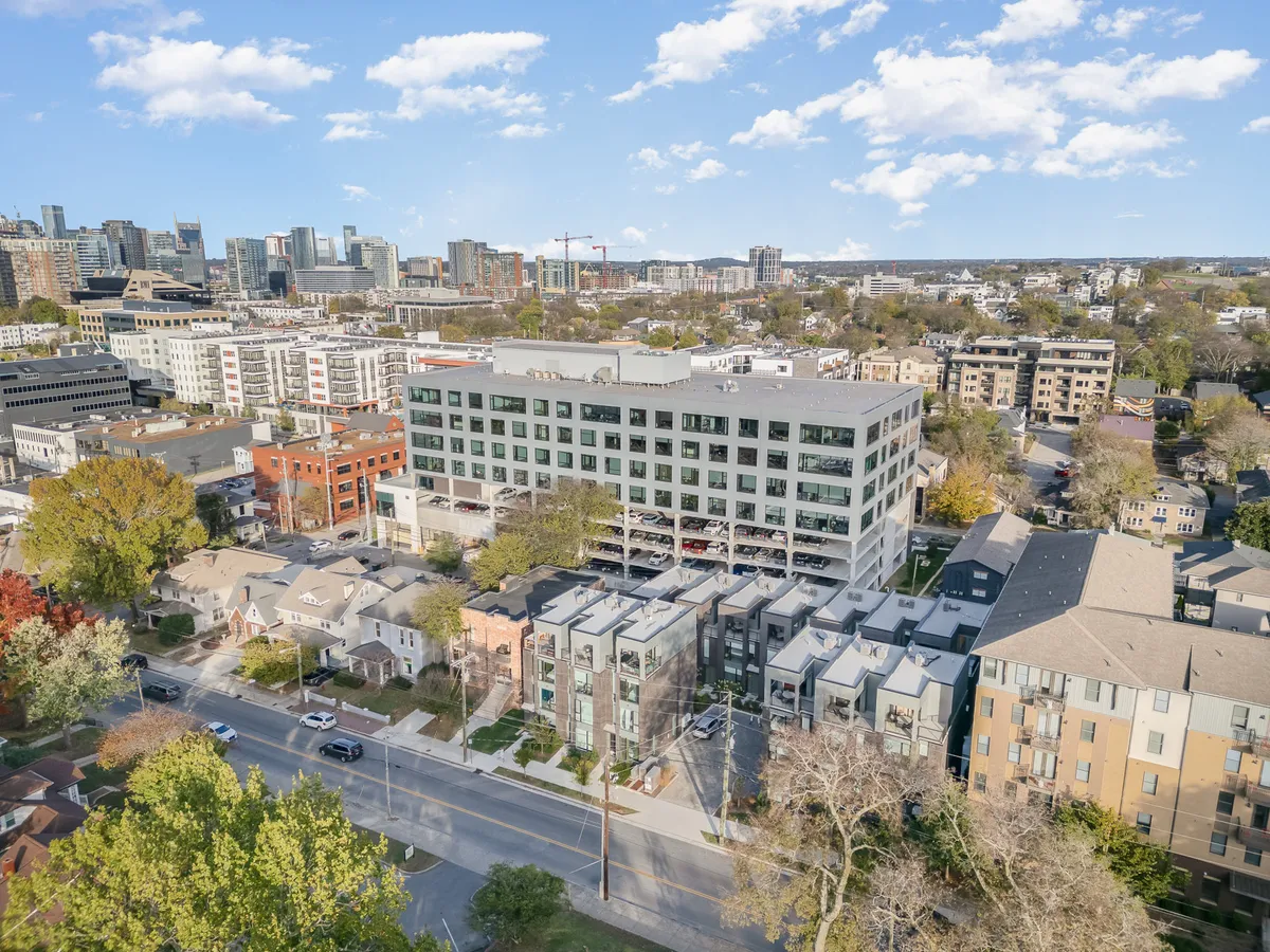 A contemporary exterior shot featuring a modern apartment building, surrounded by residential homes, green trees, and a distant view of the city skyline—evoking the feeling of urban living near Nashville’s entertainment highlights.