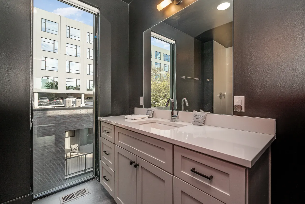 An interior image highlighting a vanity sink with a large mirror and windows that reveal glimpses of the exterior cityscape. The space is bright, functional, and contemporary, ideal for personal grooming and starting the day with city views.