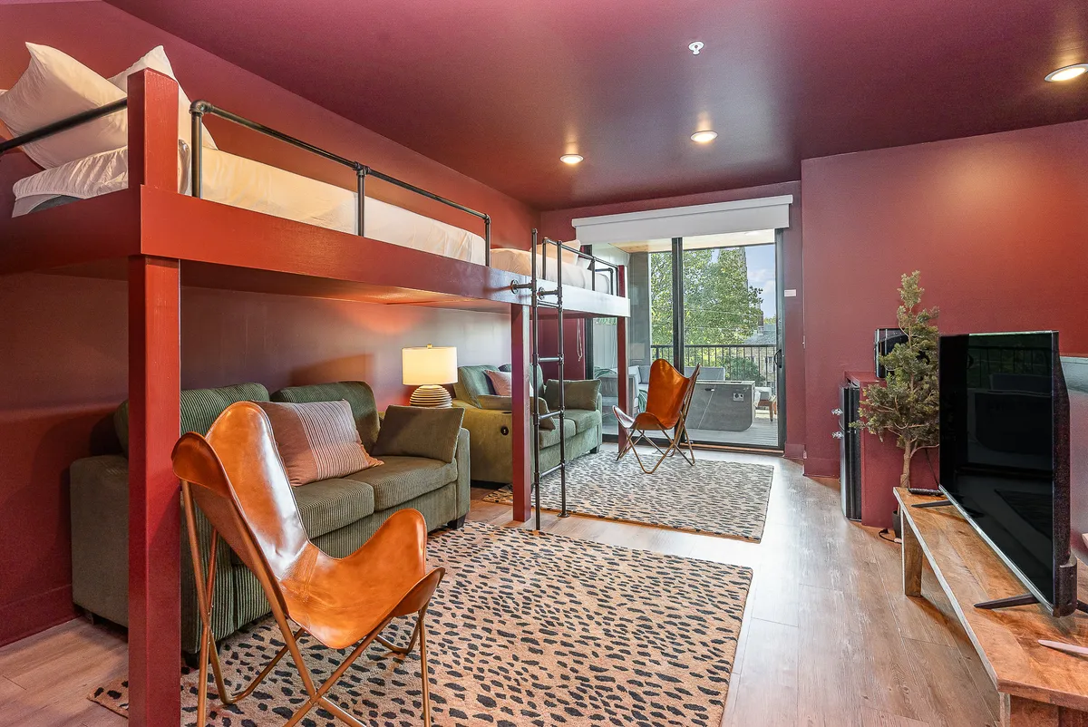 A playful yet comfortable bunk room with a queen-over-queen bunk bed, a green sofa for lounging, a leather accent chair, and a large window letting in plenty of natural light. The space feels adaptable and family-friendly, ideal for groups or children.