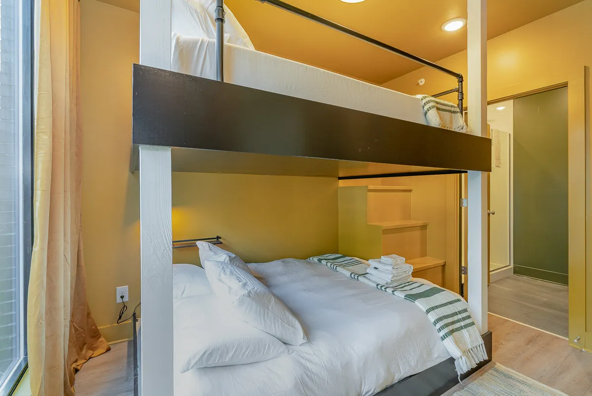A loft-style sleeping area featuring a lower and an upper bed, decorative pillows, a staircase for access, and a window with curtains. The cozy, multi-level setup is ideal for additional guests or children, evoking a sense of comfort and fun.