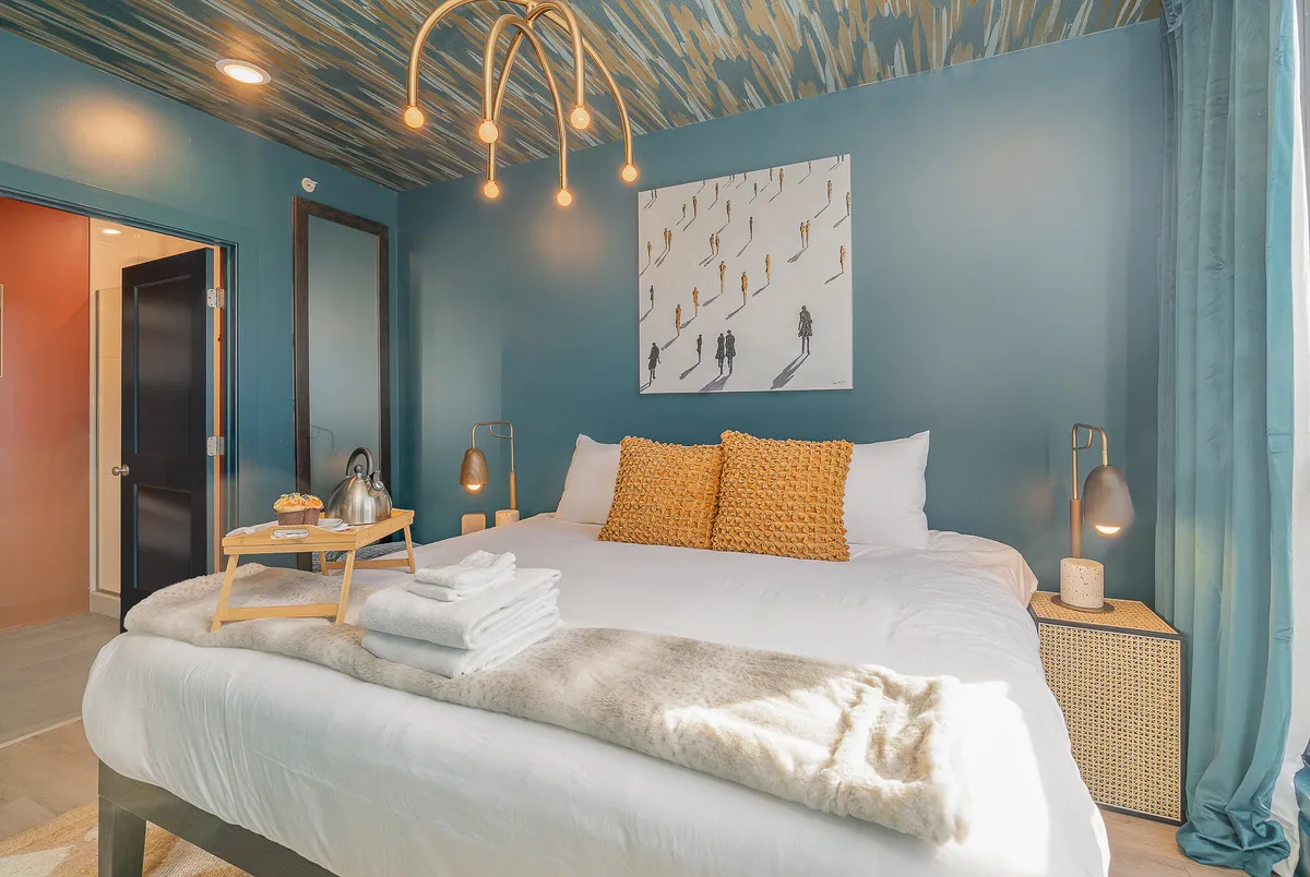 A stylish bedroom featuring a plush bed adorned with decorative pillows, soft lighting fixtures for a warm glow, tasteful artwork on the wall, and a bedside table set with a breakfast tray. Natural light streams in, enhancing the room's welcoming atmosphere.