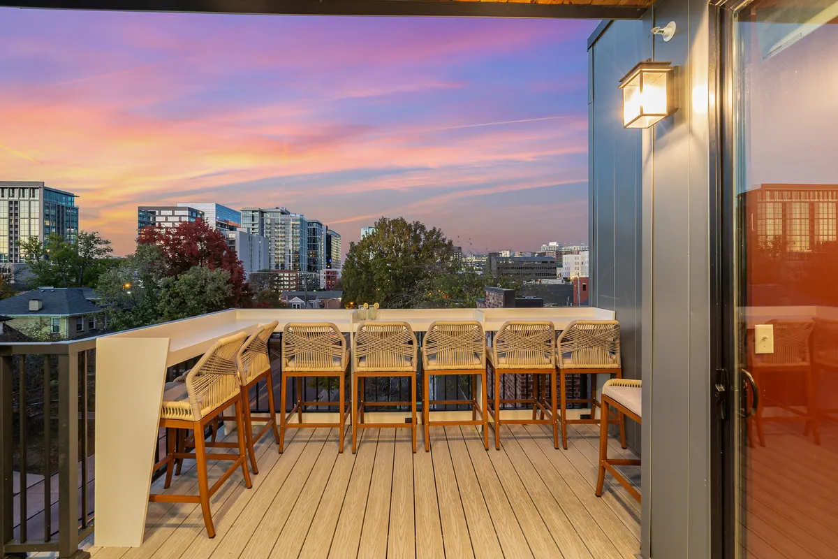 A rooftop bar area with sleek seating facing toward the city skyline, bathed in the warm colors of dusk. The open-air layout is ideal for hosting gatherings and soaking in panoramic views, while the relaxed mood invites both entertaining and tranquil moments.