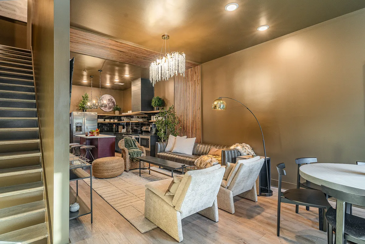 A spacious open-concept interior featuring a sparkling chandelier above a plush designer sofa, a sleek staircase, and an inviting dining table set for a communal meal. The arrangement suggests an area designed for social gatherings and memorable family meals, with contemporary decor and cozy ambiance.
