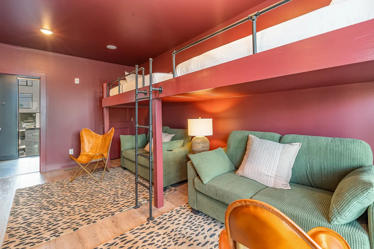 A vibrant loft area featuring a lofted bed, green sofas, an orange accent chair, a table lamp, and an animal print rug. The space feels open and flexible, with ample seating for group activities or relaxation, and playful decor that appeals to both teens and adults.